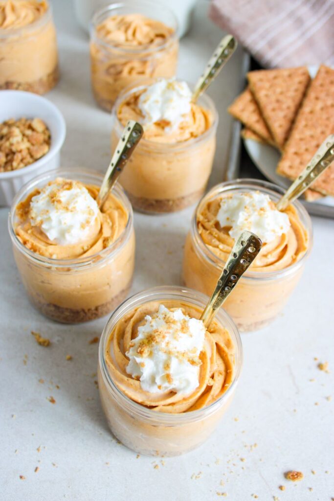 Five small glass jars filled with creamy pumpkin mousse topped with whipped cream and sprinkled with crumbled graham crackers. Each jar has a gold spoon. The jars are arranged on a light-colored surface alongside a plate of whole graham crackers and a small bowl of crumbs.