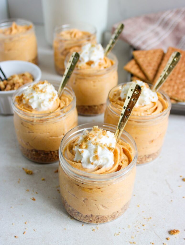 Six glass jars filled with pumpkin cheesecake mousse and topped with whipped cream. Each jar has a gold spoon and rests on a sprinkle of crushed graham crackers. In the background, there are graham crackers on a cloth and a bowl with more crumbles.