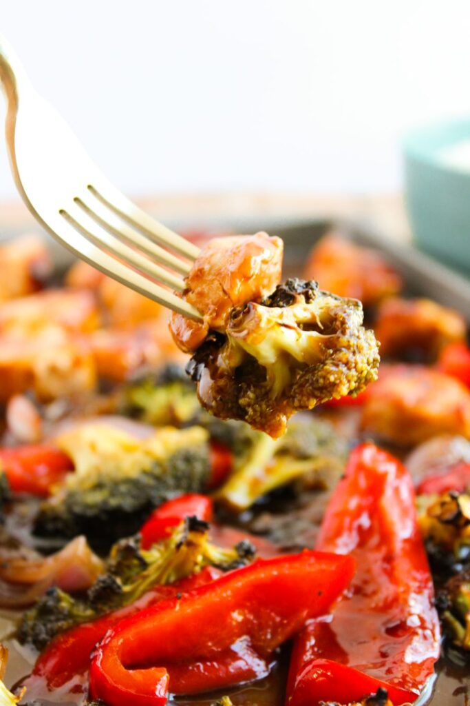 A fork holding a piece of roasted broccoli is above a dish featuring roasted vegetables, including red bell peppers and onions, with a sauce. The background is blurred to emphasize the fork and vegetables.