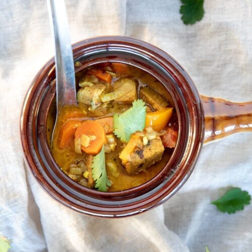 A brown ceramic bowl filled with a hearty stew, including chunks of meat, carrots, and topped with cilantro. A spoon rests inside the bowl. The bowl is on a light fabric with scattered cilantro leaves.