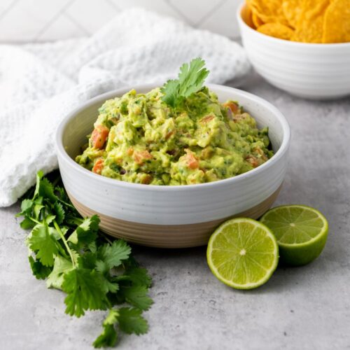 A bowl of guacamole garnished with cilantro is on a gray surface. Beside it are fresh cilantro sprigs and sliced lime. In the background, tortilla chips are visible in a separate bowl. A white cloth is casually placed nearby.