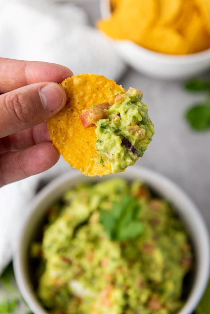 A hand holding a tortilla chip with guacamole over a bowl filled with guacamole. There are chopped vegetables visible in the dip, and a bowl of chips is in the background.