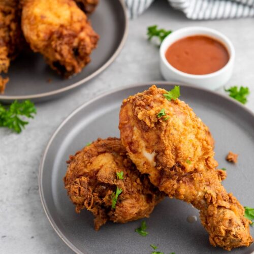 Two plates each with pieces of crispy fried chicken garnished with parsley, accompanied by a small bowl of dipping sauce on a light-colored surface.