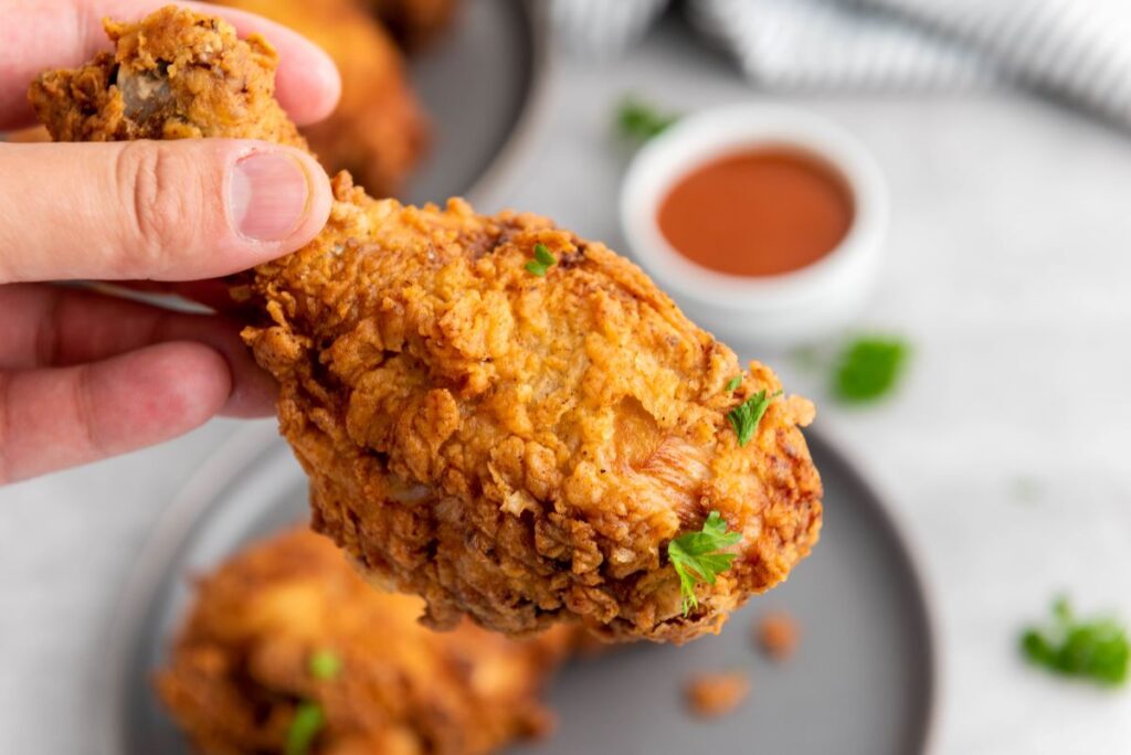 A hand holding a piece of crispy fried chicken garnished with parsley, with a plate of chicken and a small bowl of dipping sauce in the background.