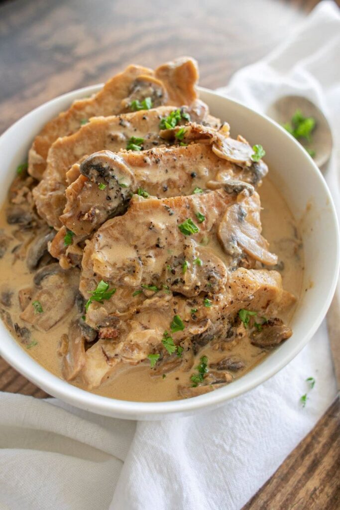 A bowl of creamy mushroom pork chops garnished with parsley. The dish features tender pork slices smothered in a rich, creamy sauce with sliced mushrooms, all presented on a wooden table with a white cloth underneath the bowl.