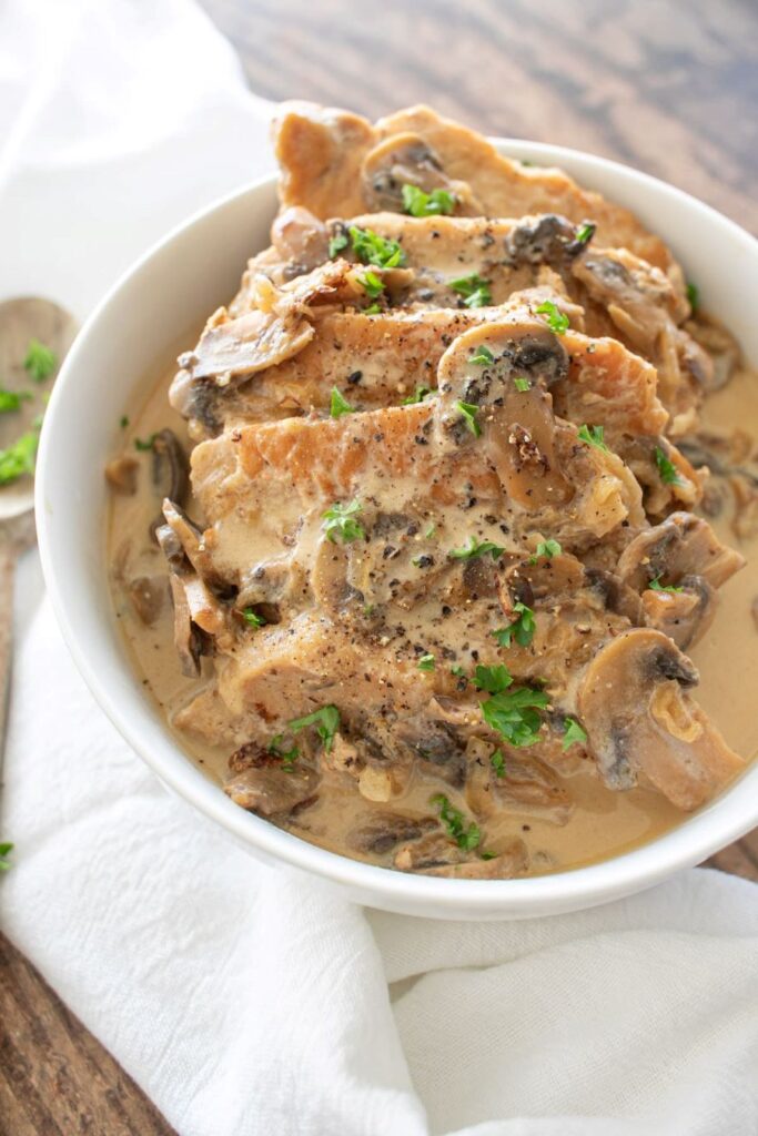 A bowl of creamy mushroom pork chops garnished with fresh parsley, sitting on a white cloth.