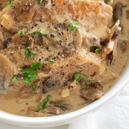 A bowl of creamy mushroom sauce with pieces of meat. The dish is garnished with freshly chopped parsley and cracked black pepper. The sauce is rich and smooth, with visible slices of mushrooms throughout.
