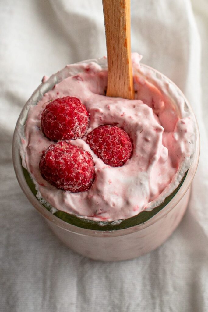 A glass jar filled with creamy pink yogurt, topped with three fresh raspberries. A wooden spoon is sticking out from the yogurt, all set against a soft, white fabric background.
