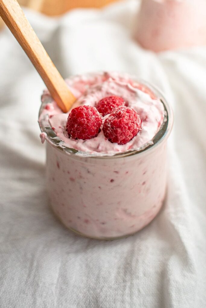 A jar of creamy raspberry mousse topped with three fresh raspberries and a wooden spoon, placed on a white textured cloth.