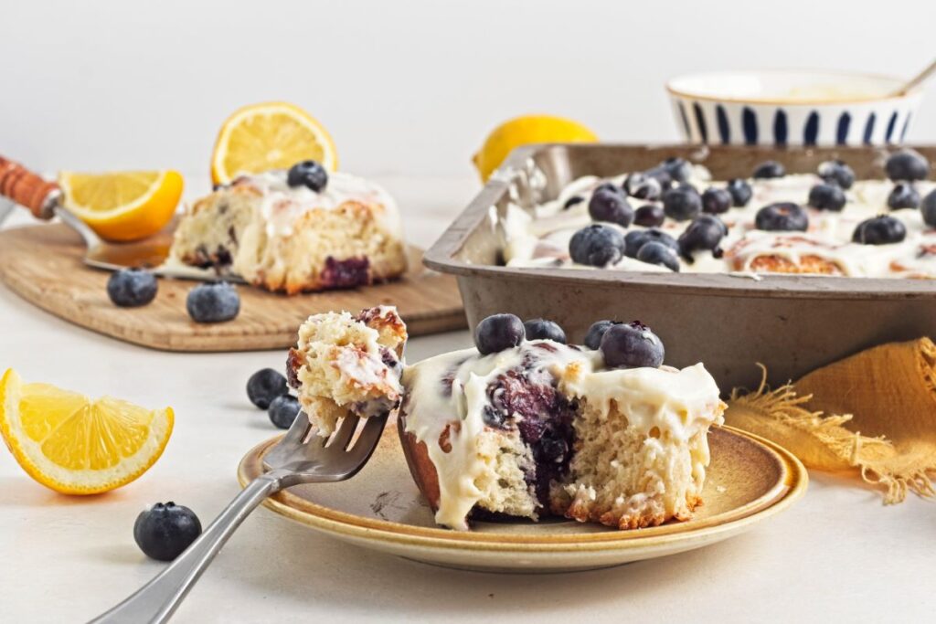 A plate with a blueberry cinnamon roll topped with white icing and fresh blueberries. A fork holds a bite. Nearby, a baking tray has more rolls, and there are lemon wedges and blueberries around on the table.