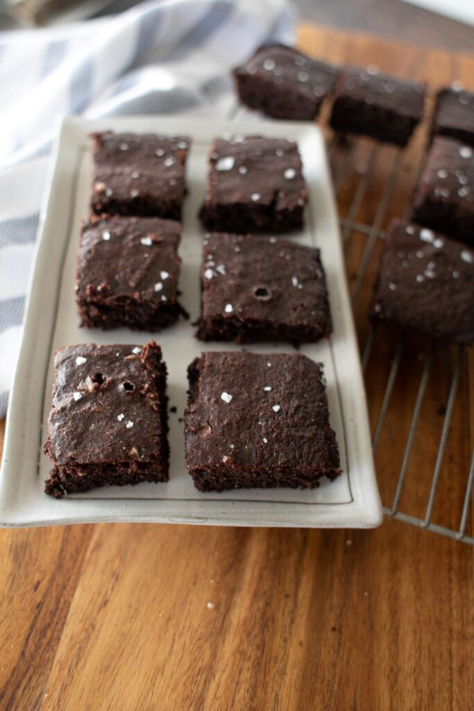 A white platter with eight square chocolate brownies sprinkled with sea salt is on a wooden table. A cooling rack with more brownies and a striped cloth are partially visible in the background.