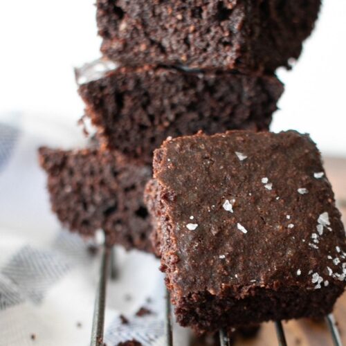 Close-up of a stack of rich, dark chocolate brownies on a cooling rack. The top brownie is sprinkled with coarse sea salt. A striped kitchen towel is visible in the background.