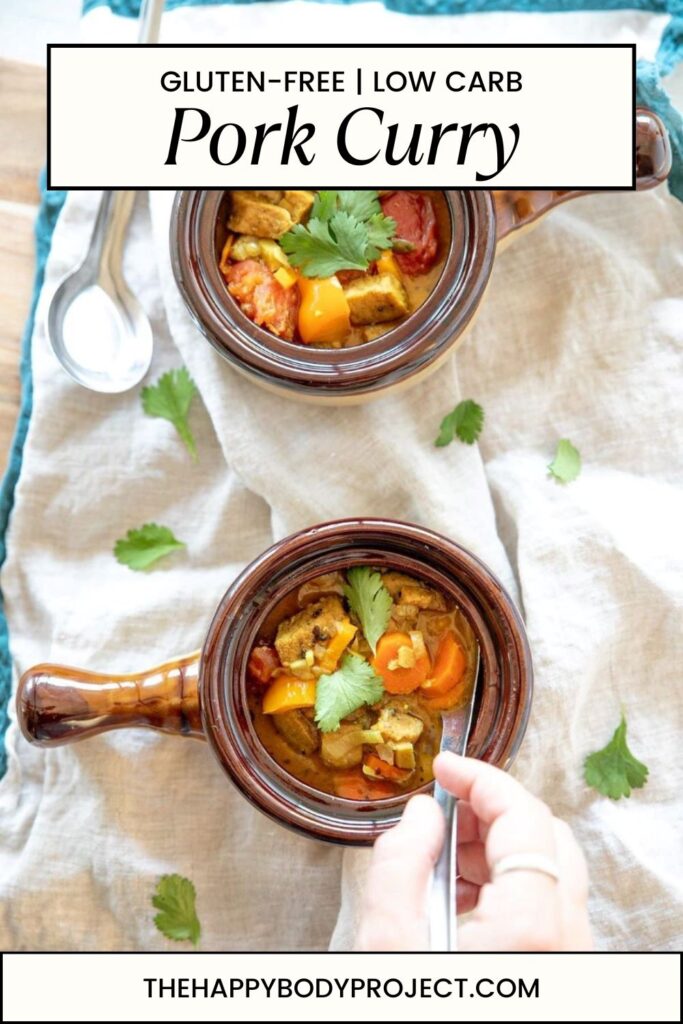 Two bowls of pork curry garnished with cilantro are on a table, accompanied by spoons. Text on the image reads "Gluten-Free | Low Carb Pork Curry" and "thehappybodyproject.com." A person's hand is holding one of the bowls.