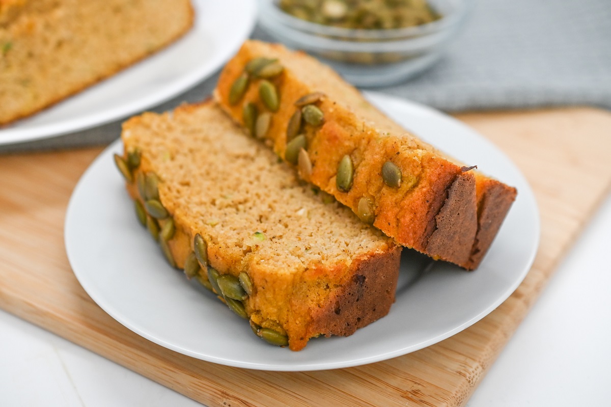 Two slices of pumpkin bread with pumpkin seeds on top are served on a white plate. A small bowl of seeds is in the background, all set on a wooden cutting board.