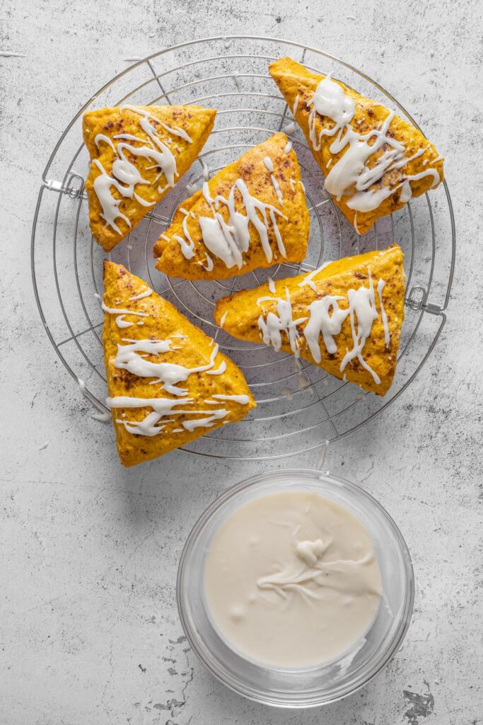 Five pumpkin scones drizzled with icing are placed on a round wire rack. Below the rack, there is a bowl filled with white icing on a light gray textured surface.