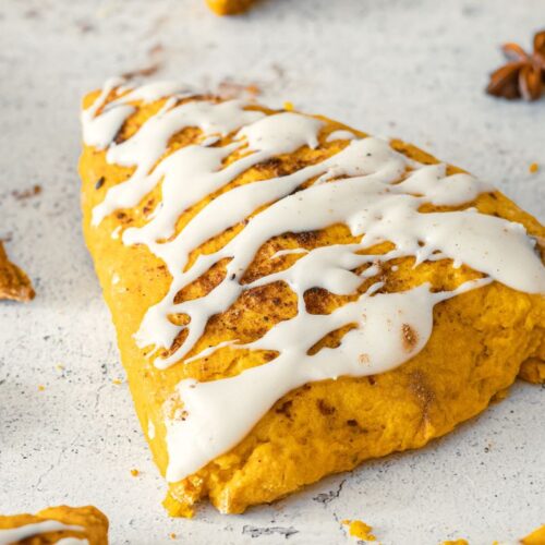 A close-up of a pumpkin scone topped with white icing drizzle on a light, textured surface. The scone is surrounded by star anise and a cinnamon stick, adding a warm, autumnal touch.