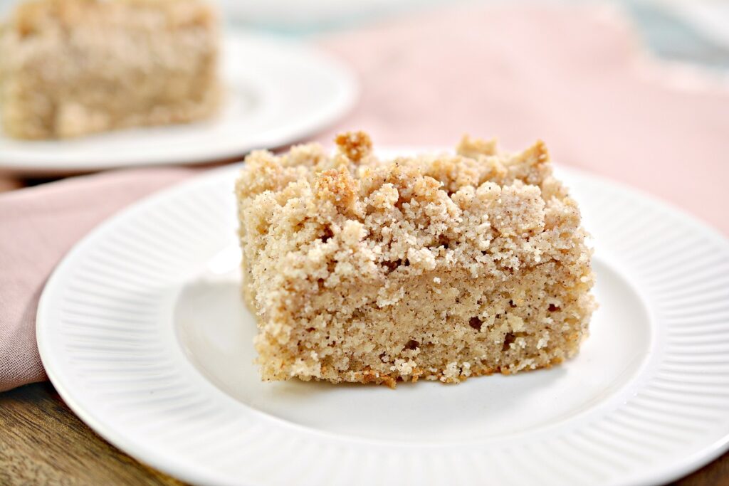A slice of crumb cake on a white plate, showcasing a crumbly topping and moist interior. Another slice is visible in the background, slightly blurred. The scene is set against a soft pink cloth.