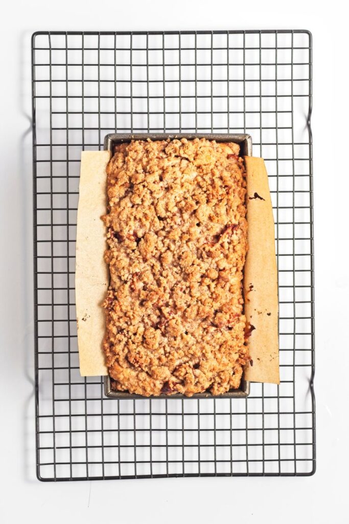 A freshly baked loaf with a crumbly, golden-brown topping sits in a parchment-lined rectangular pan on a wire cooling rack against a white background.