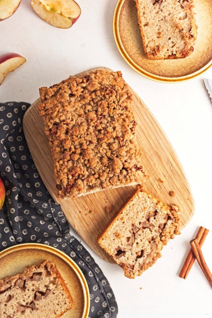 A loaf of apple cinnamon bread with a crumbly topping on a wooden board, partially sliced. Two additional slices are on small plates nearby. Cinnamon sticks and apple slices are scattered around, with a dark napkin in the corner.