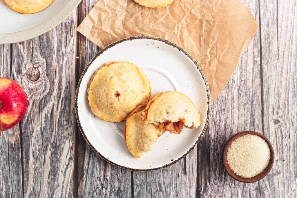 A plate with two apple hand pies, one partially eaten, on a rustic wooden table. An apple and a small bowl of sugar are nearby. The background includes a piece of brown parchment paper and part of another plate with more pastries.