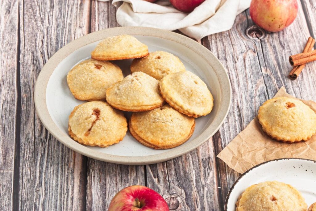 A plate of small, round, golden-brown apple pies on a wooden table. Surrounding the plate are fresh apples and cinnamon sticks. A few more pies are on a small plate and parchment paper. A beige cloth is in the background.
