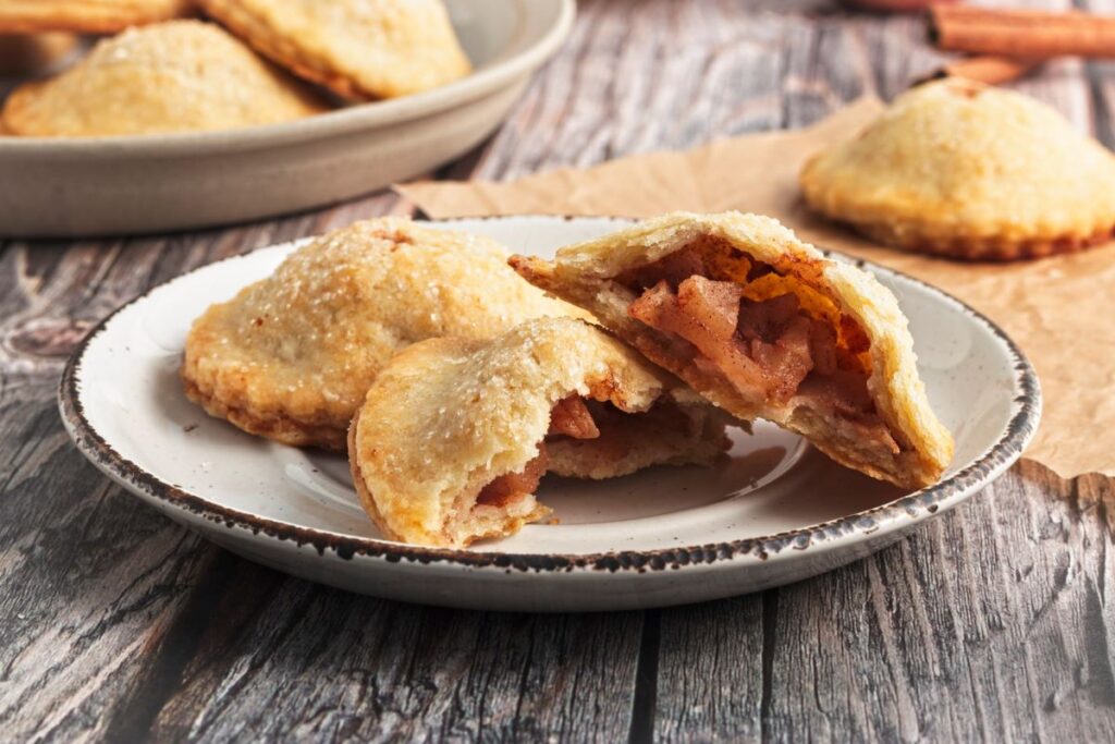 A plate of small hand pies with a flaky, golden-brown crust, filled with spiced apple chunks. One pie is cut open to show the filling. Additional pies are in the background on a wooden table.