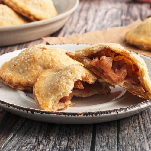 A plate of small hand pies with a flaky, golden-brown crust, filled with spiced apple chunks. One pie is cut open to show the filling. Additional pies are in the background on a wooden table.