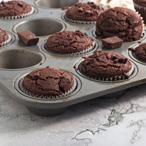 A muffin tray with six chocolate muffins and empty slots sits on a marble surface. Two muffins are on a wooden board, one on a white plate.