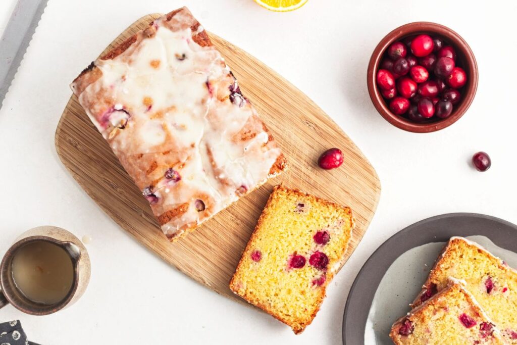 Loaf of cranberry-orange bread with icing on a wooden cutting board. A slice is cut and placed nearby. Fresh cranberries in a bowl and additional slices on a plate. A cup of coffee is in the corner.
