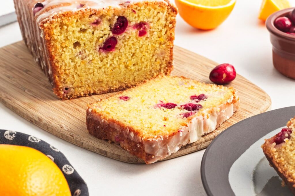 A sliced loaf of orange cranberry bread topped with glaze sits on a wooden cutting board. Fresh cranberries and orange slices are placed around it, along with a gray plate holding another slice of the bread.