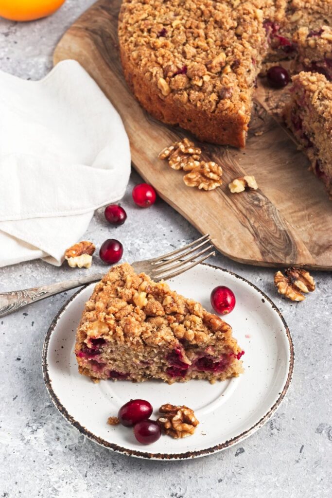 A slice of cranberry walnut crumb cake on a white plate with scattered cranberries and walnuts. The whole cake is on a wooden board in the background, partially sliced, next to a white cloth. A fork lies beside the plate.