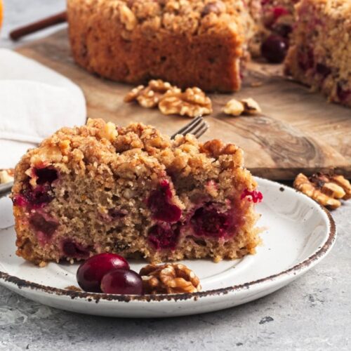 A slice of cranberry cake with a crumbly topping on a white plate, garnished with fresh cranberries and walnuts. The background shows the remaining cake on a wooden board, a white napkin, and part of an orange.