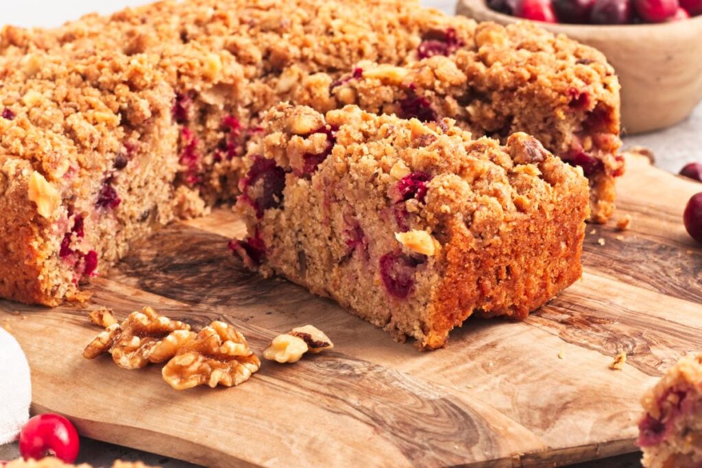 A close-up of a slice of cranberry walnut coffee cake on a wooden board. The cake is topped with crumbled streusel and surrounded by whole walnuts. A bowl of fresh cranberries is blurred in the background.