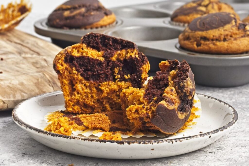 A close-up of a marbled chocolate and pumpkin muffin on a white plate, broken open to show the texture. A muffin tin with more muffins is in the background. The setting is on a gray surface with a wooden board nearby.