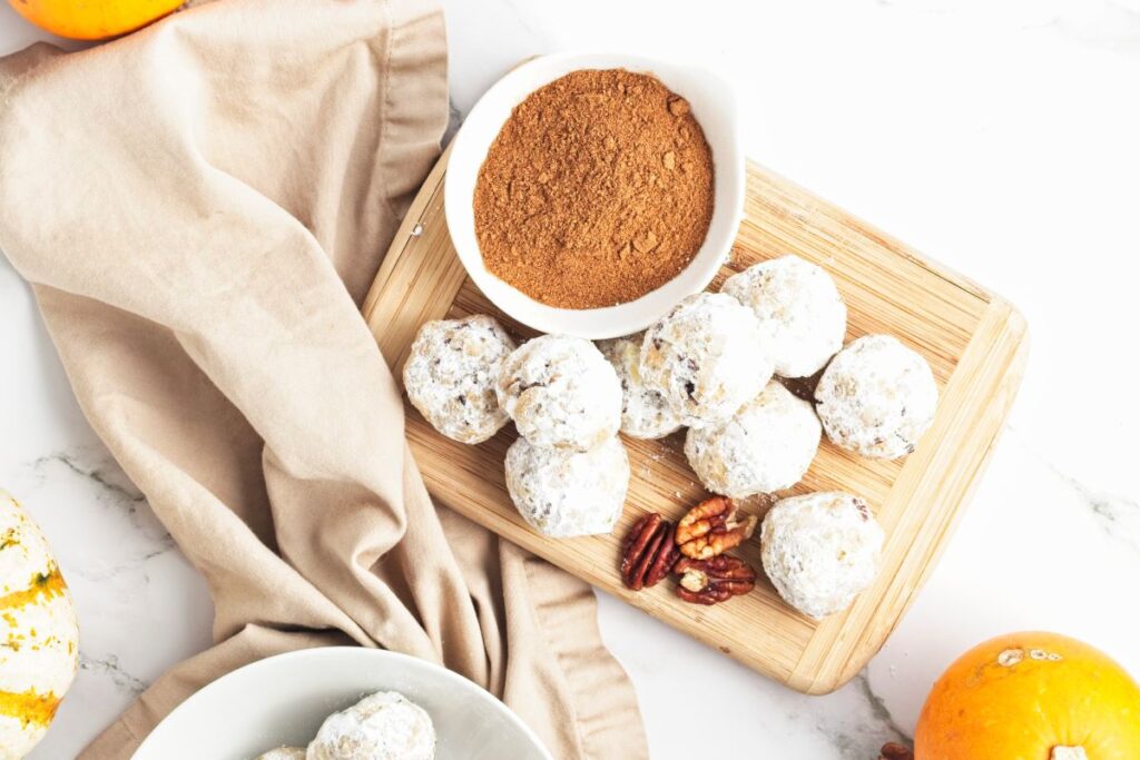 A wooden cutting board with powdered sugar-coated bite-sized snacks, a small bowl of cinnamon, pecans, and an orange. A beige cloth is draped next to the board on a white marble surface, with additional snacks in a bowl nearby.