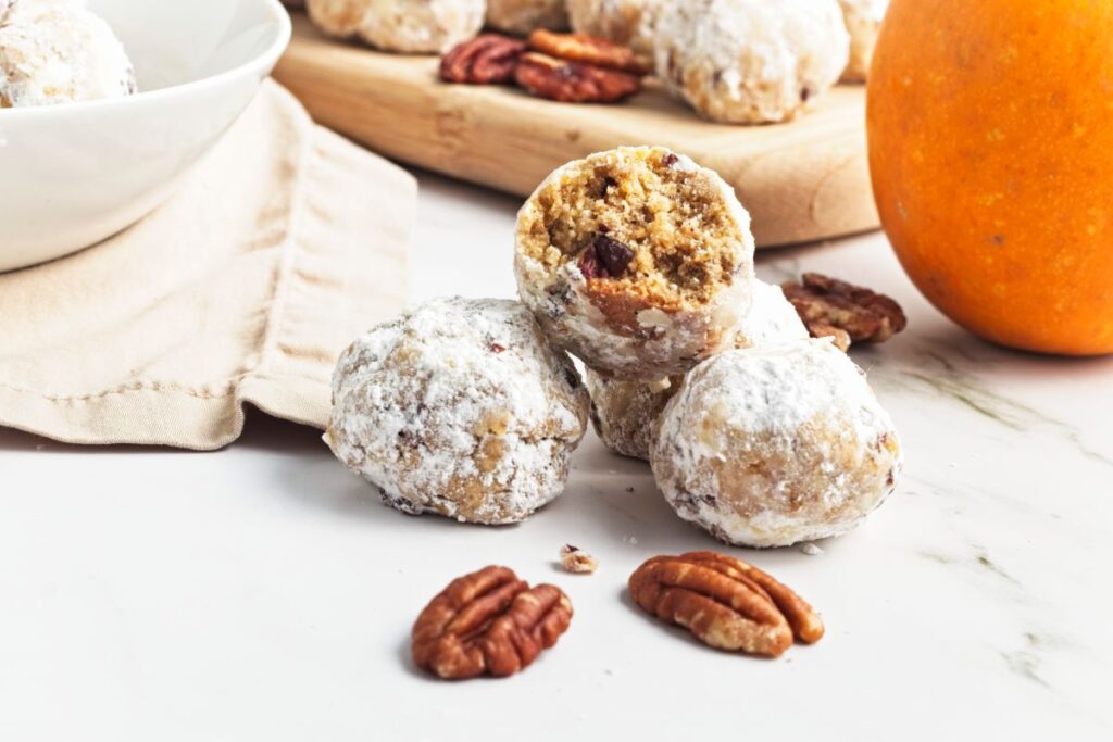 A close-up of three powdered sugar-coated cookies on a white surface, with one partially eaten cookie revealing a nutty filling. There are a few pecans scattered around, a white bowl partially visible on the left, and an orange pumpkin on the right in the background.