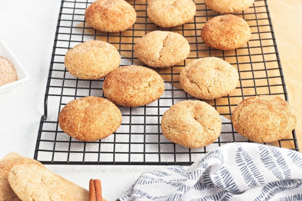Freshly baked snickerdoodle cookies cooling on a black wire rack. Some cookies are slightly cracked, with a cinnamon and sugar coating. A striped cloth and a cinnamon stick are nearby on a light countertop.