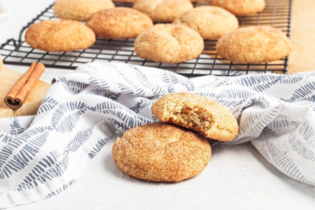 Snickerdoodle cookies on a cooling rack and linen cloth. One cookie in the foreground is broken, revealing a soft interior. A cinnamon stick is nearby, and the cookies have a cinnamon-sugar coating.