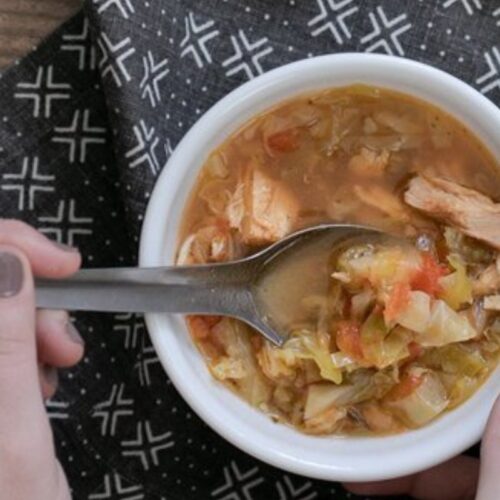 A person holding a bowl of soup with chunks of meat and vegetables, using a spoon. The bowl is on a patterned black cloth.