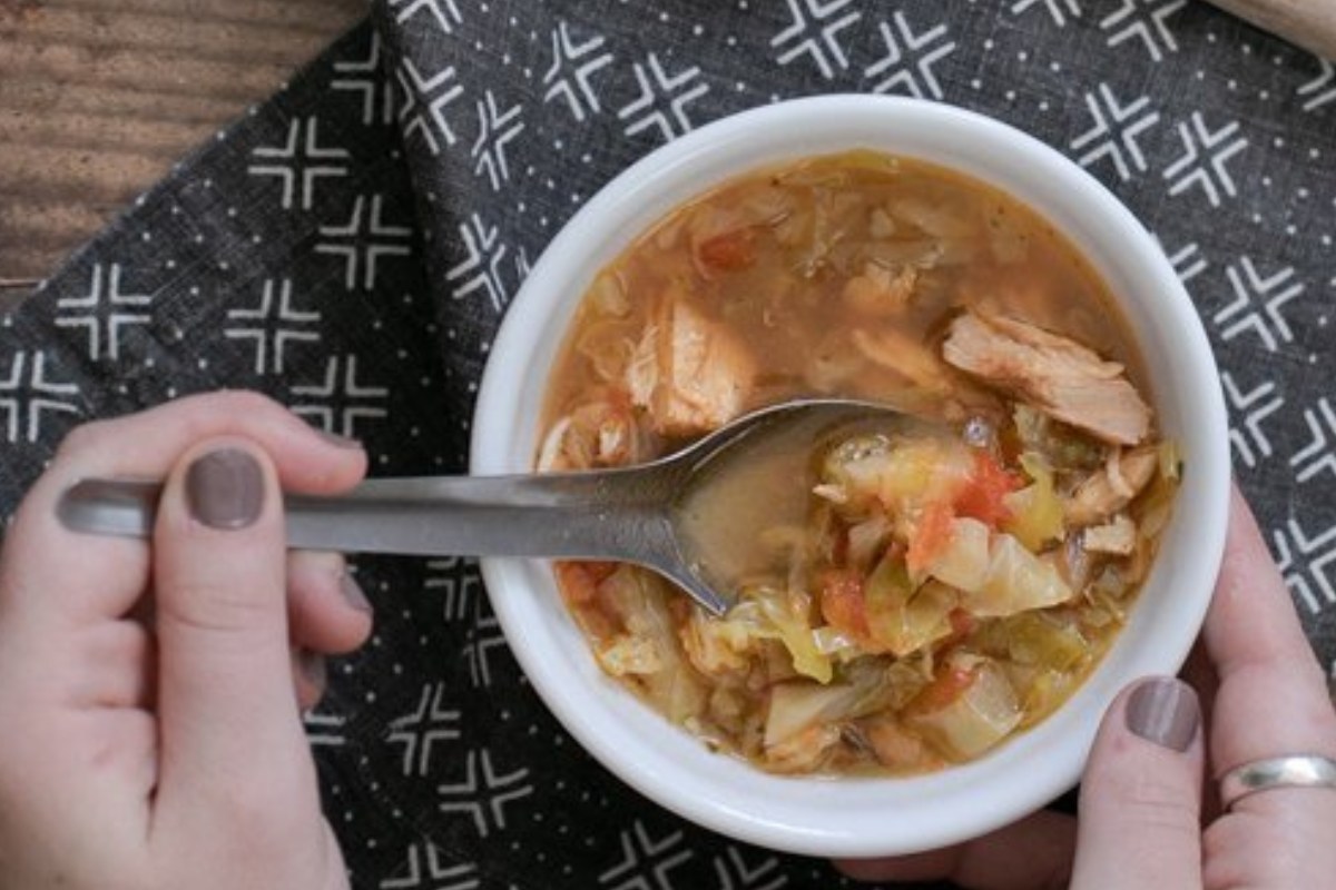 A person holding a bowl of soup with chunks of meat and vegetables, using a spoon. The bowl is on a patterned black cloth.