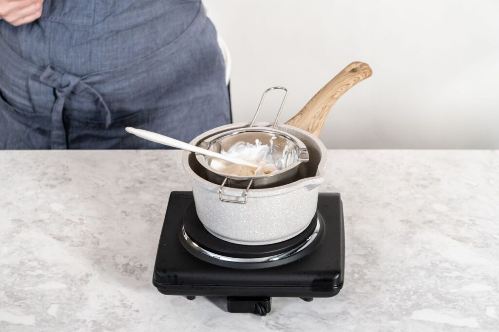 A person wearing a gray apron stands behind a pot on an electric stove. The pot contains a smaller bowl with a white mixture being stirred with a spatula. The setup is on a light-colored countertop.