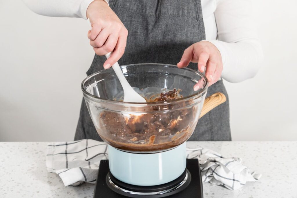 A person wearing a gray apron stirs melted chocolate in a glass bowl with a white spatula. The bowl is placed over a saucepan on a stovetop. A checkered cloth is nearby on the white countertop.