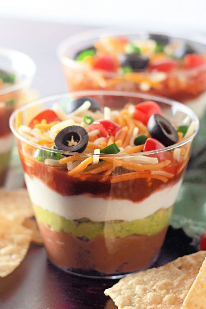 A clear plastic cup filled with layered dip featuring refried beans, guacamole, sour cream, salsa, shredded cheese, diced tomatoes, green onions, and sliced black olives. Tortilla chips are nearby.