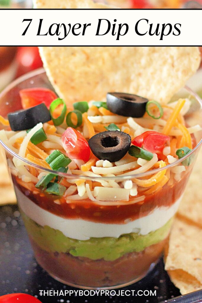A clear cup filled with seven layers of colorful dip, including guacamole, sour cream, salsa, shredded cheese, chopped tomatoes, and olives. A tortilla chip is inserted into the dip. Text at the top reads "7 Layer Dip Cups.