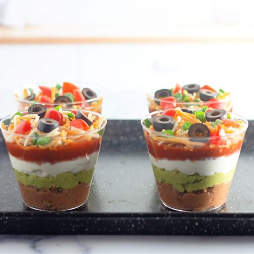 Four individual servings of layered dip in clear cups sit on a black tray. Each cup features layers of beans, guacamole, sour cream, salsa, cheese, and is topped with chopped tomatoes, green onions, and sliced black olives.