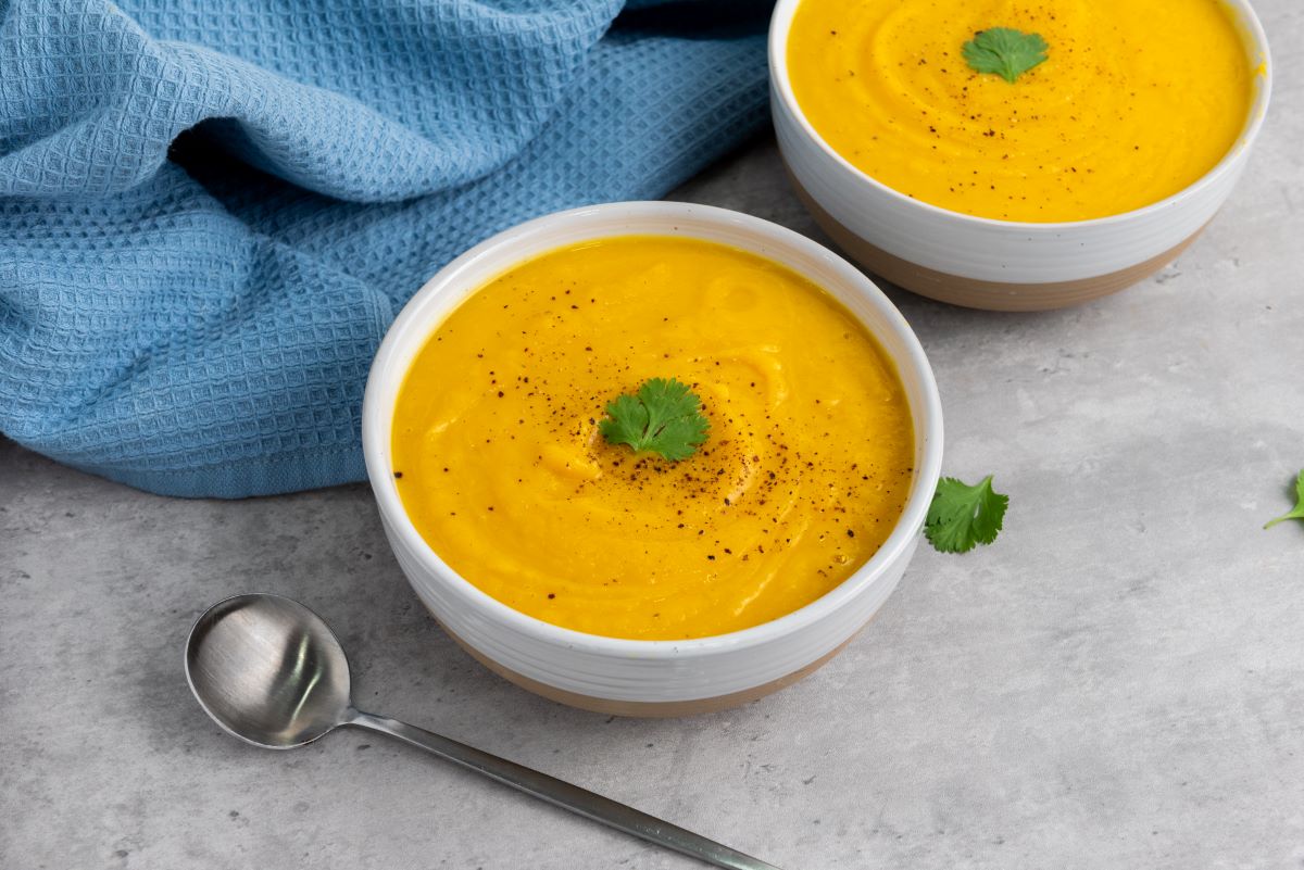 Two bowls of orange soup garnished with cilantro sit on a gray surface. A blue cloth and a spoon are placed nearby. The soup appears creamy and is lightly sprinkled with black pepper.
