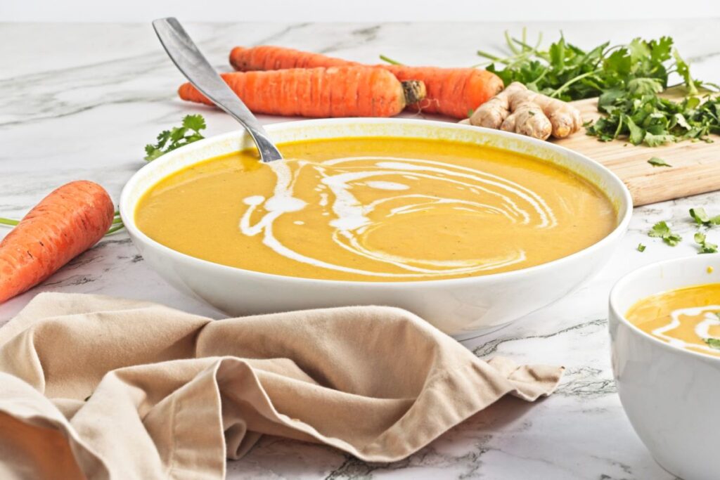A bowl of creamy carrot soup garnished with swirls of cream sits on a marble surface. Fresh carrots, ginger, and cilantro are in the background, and a beige cloth is placed nearby.