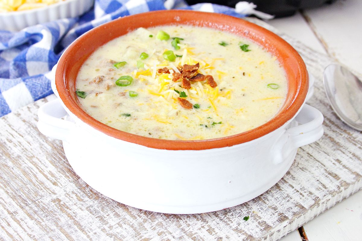 A creamy soup served in a white bowl with a brown rim, topped with shredded cheese, bacon bits, and sliced green onions. A blue and white checkered cloth is in the background.