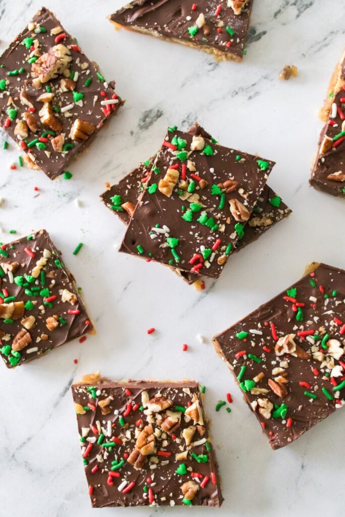 Top view of several pieces of toffee topped with chocolate, colorful sprinkles, and chopped nuts arranged on a white marble surface. The treats are cut into squares, showcasing a festive mix of red, green, and white sprinkles.