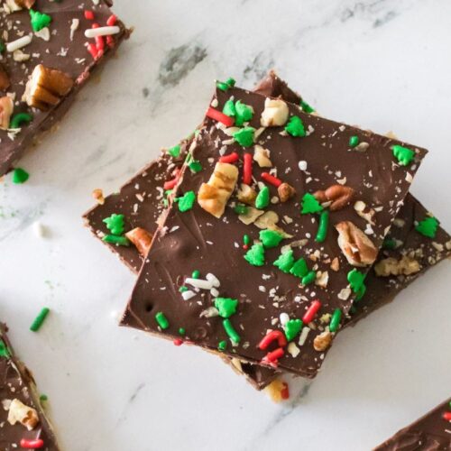 A stack of chocolate-covered toffee squares topped with festive red and green sprinkles and chopped nuts on a white marble surface. More pieces are scattered around.